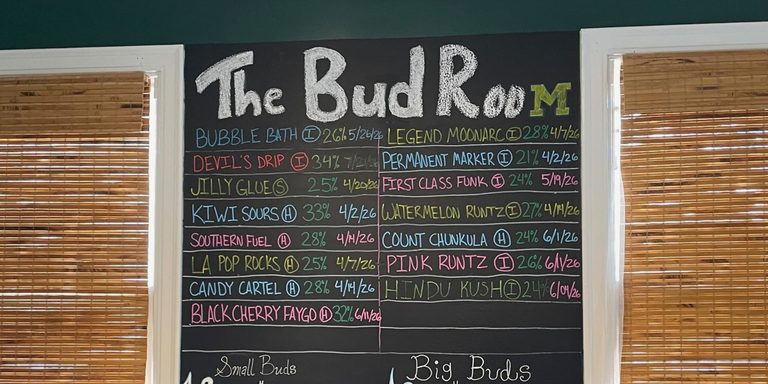 A chalkboard sign reading The Bud Room with several cannabis strains listed for sale.