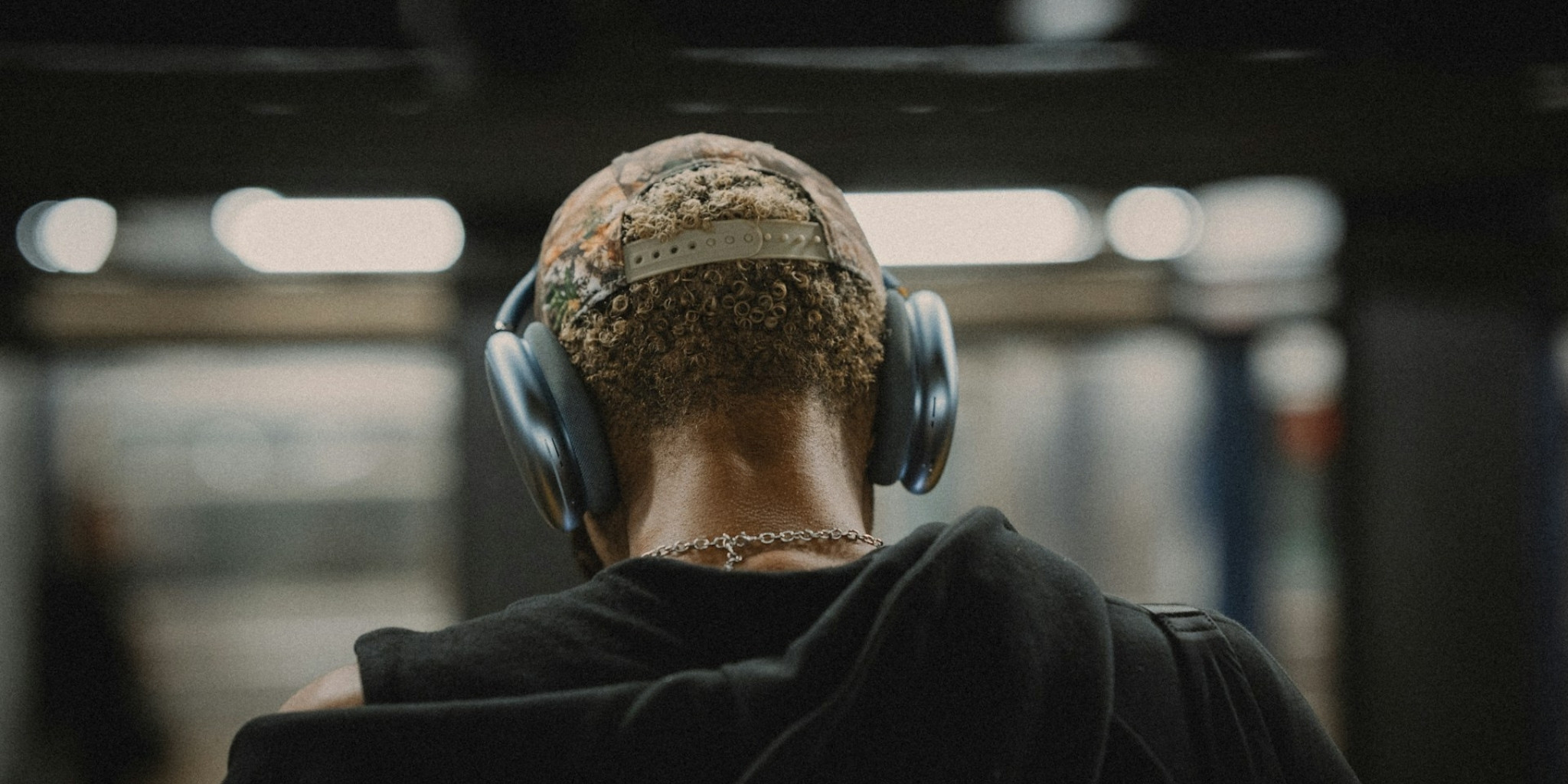 The back of a mans head with headphones on standing on a train platform